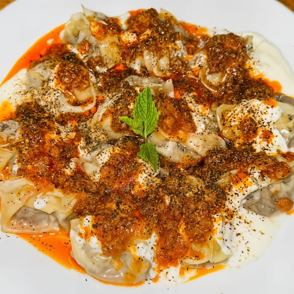 Afghan Mantu dumplings topped with yogurt, spicy sauce, dried herbs, and a mint sprig.